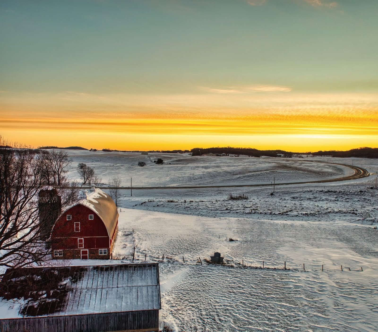 Aerial view of a red barn and a winding road across snowy farmland at sunset