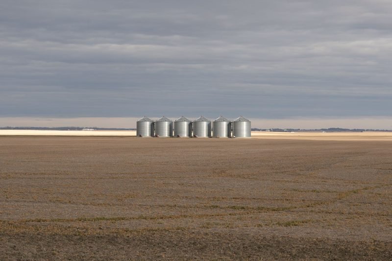 A line of small grain bins far off across flat brown land under grey cloud
