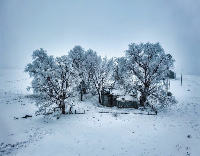 A farmhouse behind frost-covered trees on open snowy ground