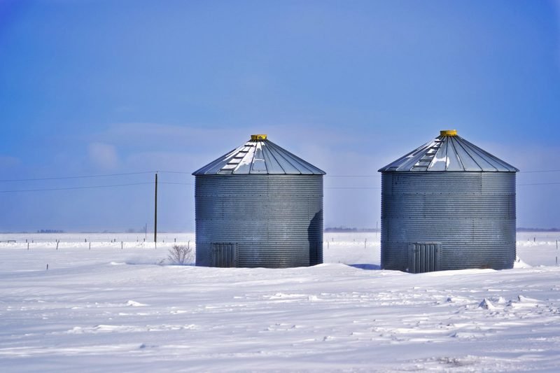 Two steel grain bins standing in deep snow under a blue sky