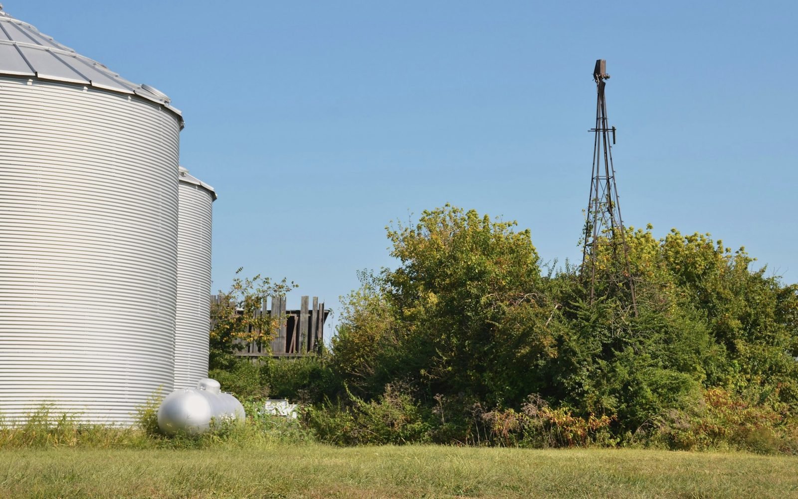 A small white propane tank at the foot of two grain bins beside an old windmill